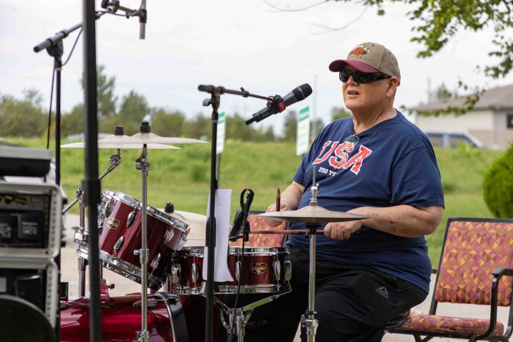 A drummer in a blue USA-themed t-shirt playing a maroon drum set outdoors.