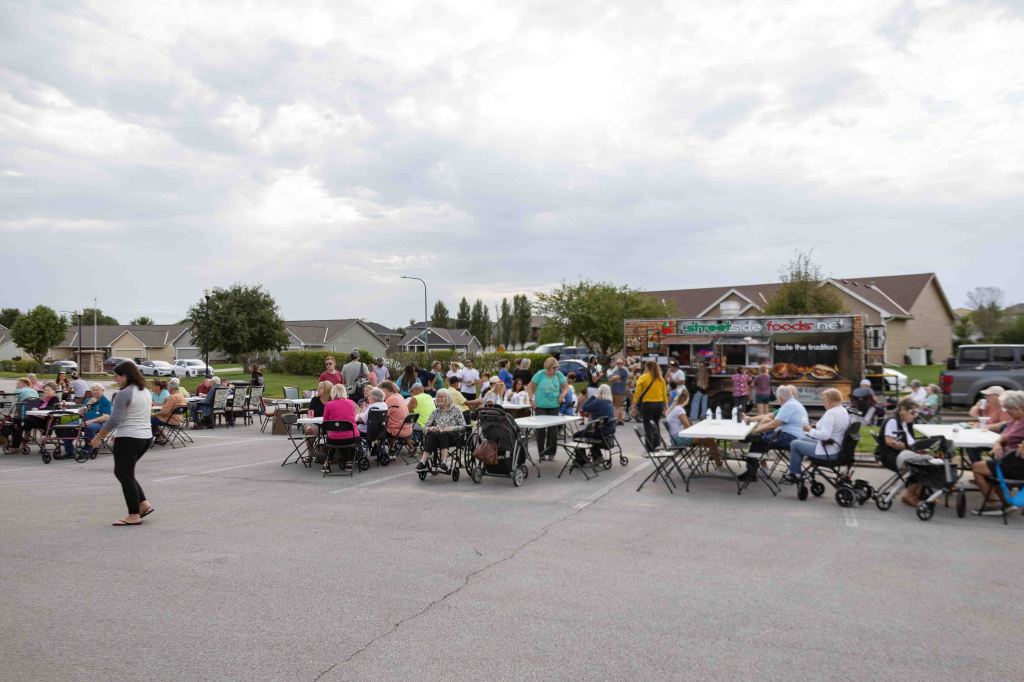 A community gathering with many people sitting at tables outdoors, enjoying food from a food truck, on a cloudy day.