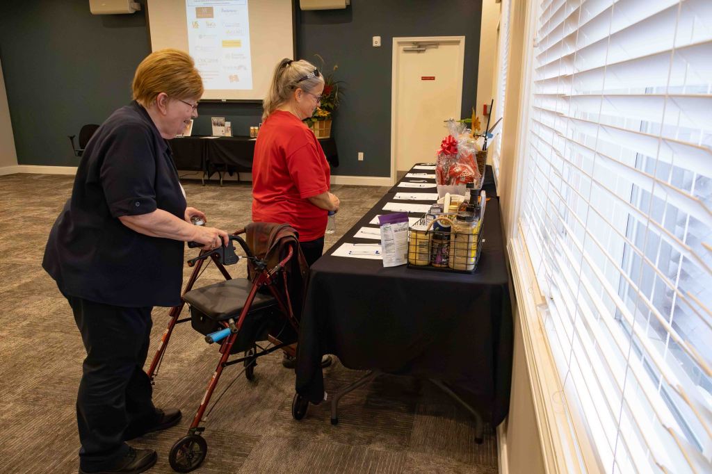 Two women interacting near a display table in a community space, one using a walker and the other standing beside her.