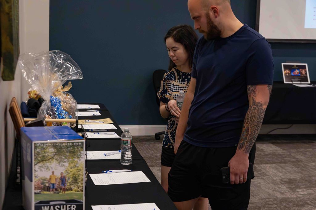 Two individuals examining a table with documents and items for a raffle, including a gift basket and a box labeled 'WASHER'.