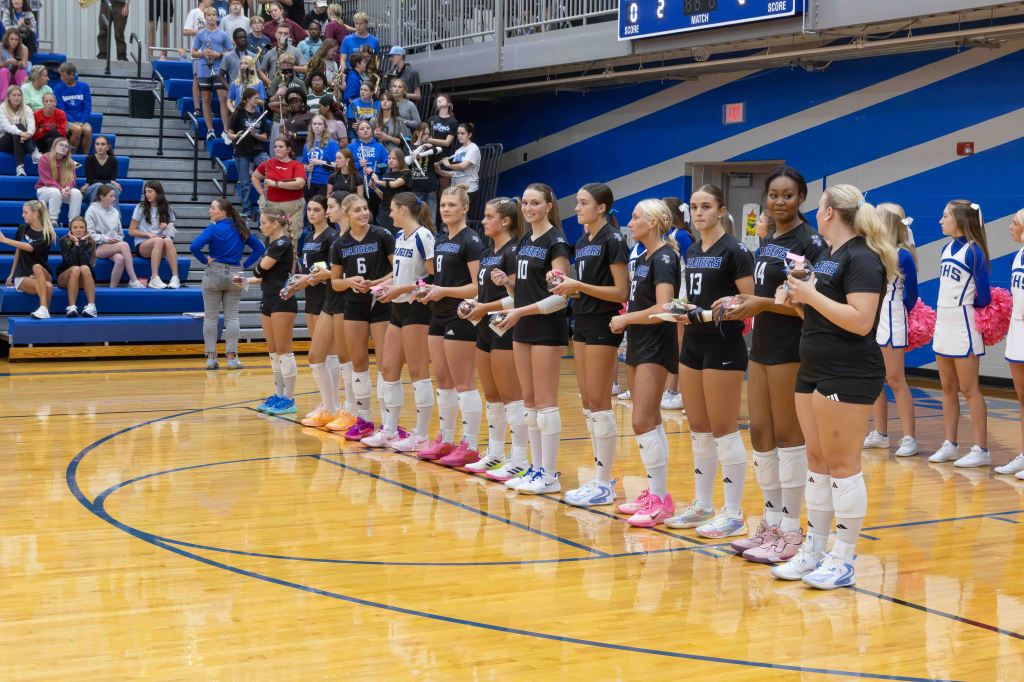 A volleyball team stands in line on the court during an event, wearing black uniforms and holding participation awards, with a crowd of spectators visible in the background.