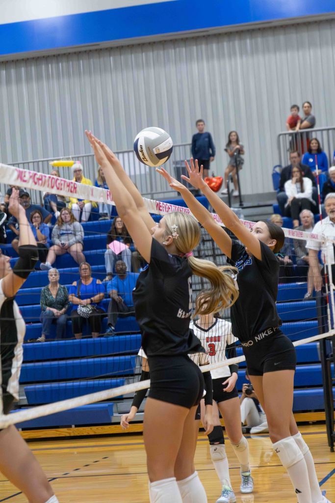 Two female volleyball players jumping to block a volleyball at the net during a match, with spectators in the background.