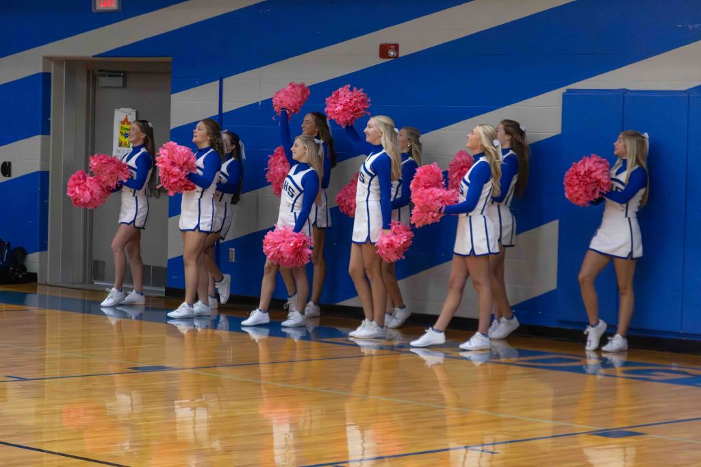 A group of cheerleaders in blue and white uniforms, holding pink pom-poms, standing along a gym wall.