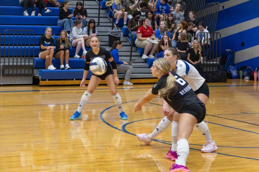 A volleyball match in a gymnasium, featuring players receiving a serve. One player in a black uniform is preparing to pass the ball, while another in a white uniform is positioned behind her. In the background, a group of spectators is watching the game.