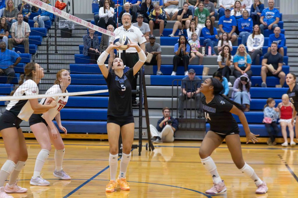 A volleyball match in progress with players in black and white uniforms on the court. One player is setting the ball while teammates prepare for a play. The bleachers are filled with spectators.