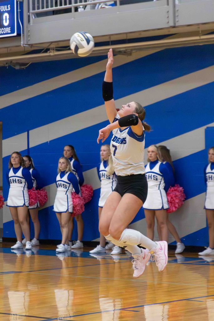 A volleyball player in mid-jump serving the ball, surrounded by cheerleaders in blue and white uniforms.