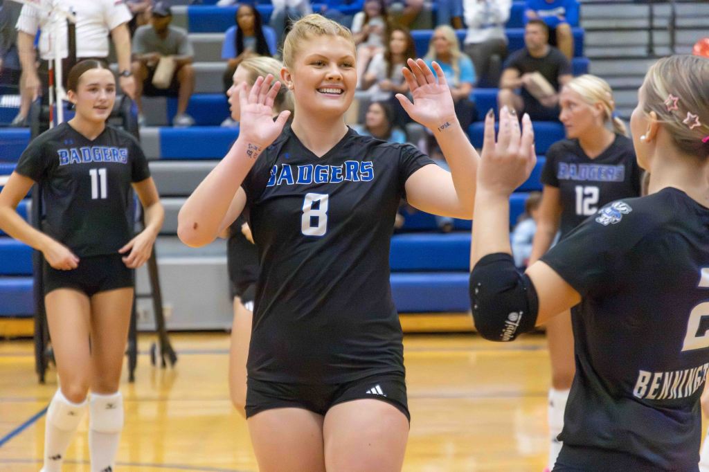 A volleyball player wearing a black jersey with "Badgers" printed on it, smiling and gesturing with hands raised during a match. Other teammates are visible in the background.