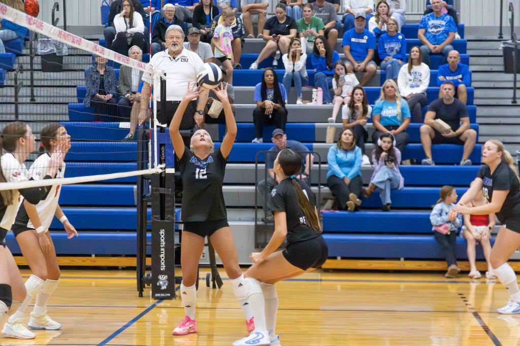 A volleyball player in a black uniform is preparing to set the ball during a game, while teammates and spectators observe in a gymnasium.
