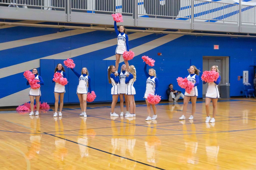 A cheerleading team performs in a gym, with one cheerleader elevated above the others while holding pom-poms. The athletes wear blue and white uniforms, and the background features blue walls and hardwood flooring.