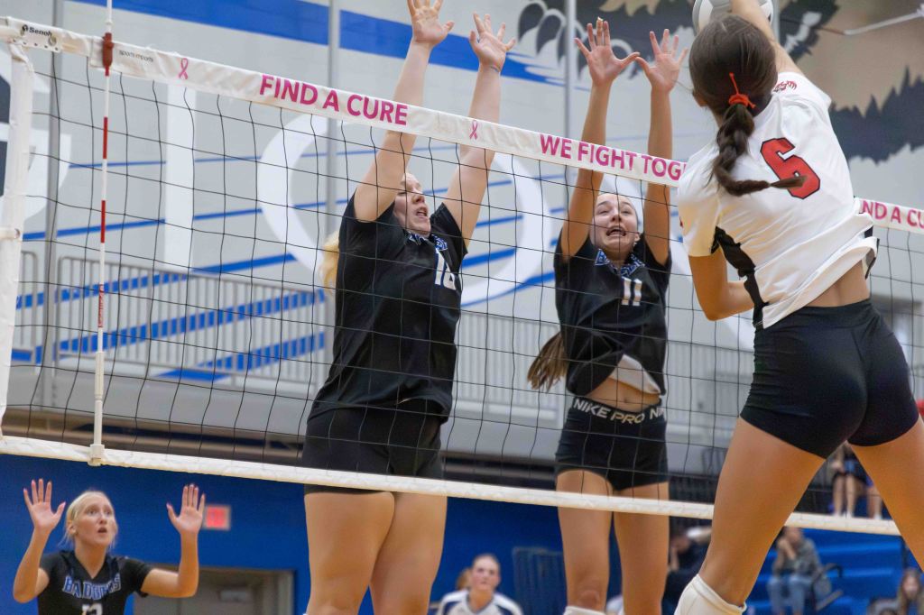 A volleyball game in progress, featuring a player in a white jersey attempting to spike the ball over the net, while two defenders in black jerseys jump to block the shot. One player in the background raises her arms in anticipation.