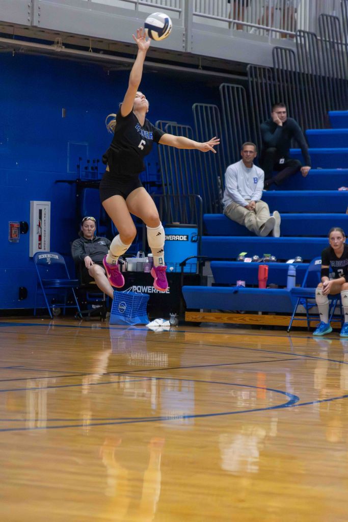 A volleyball player in a black uniform jumps to serve the ball, with spectators in the background.