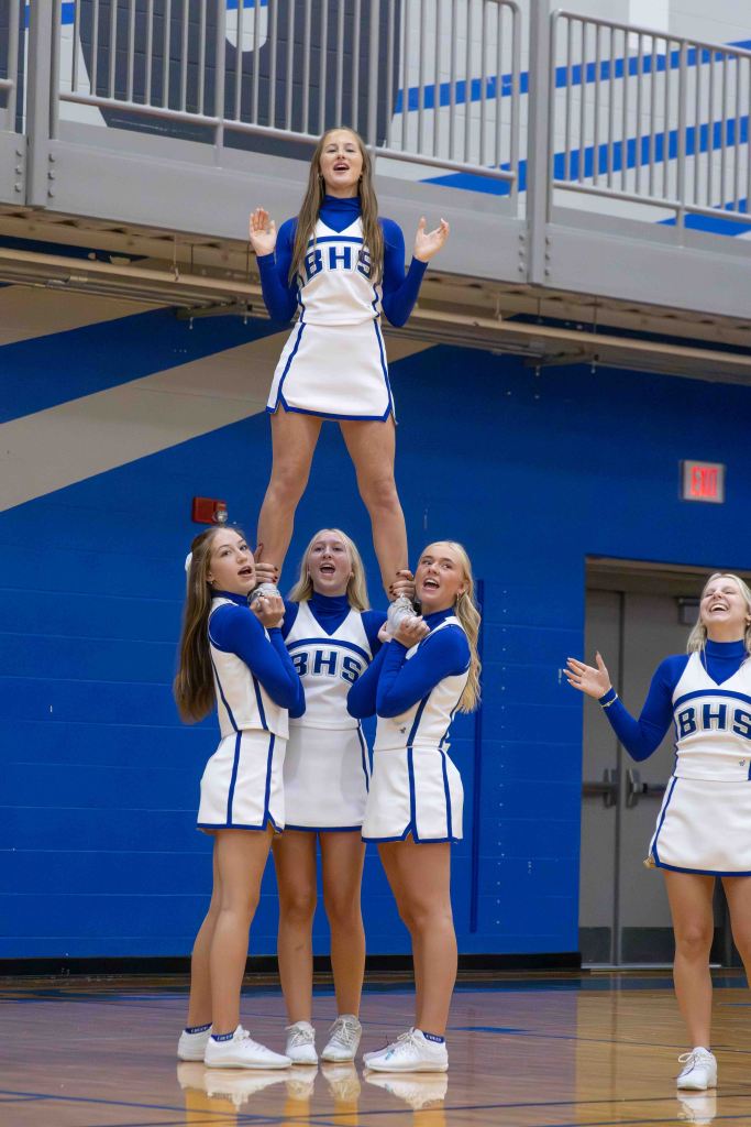 A cheerleading squad performing a stunt, featuring one cheerleader lifted above others in a gymnasium with blue walls.