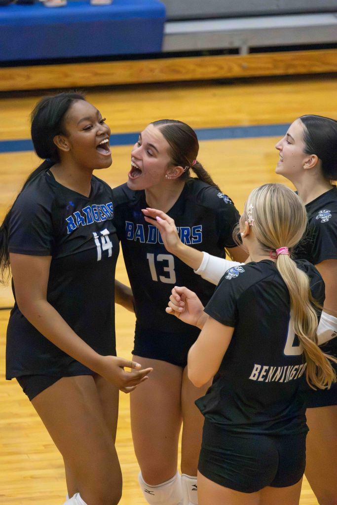 A group of four female volleyball players celebrating together on the court, smiling and showing excitement. Players are wearing black jerseys with blue accents, and one jersey has 'Badgers' written on it.
