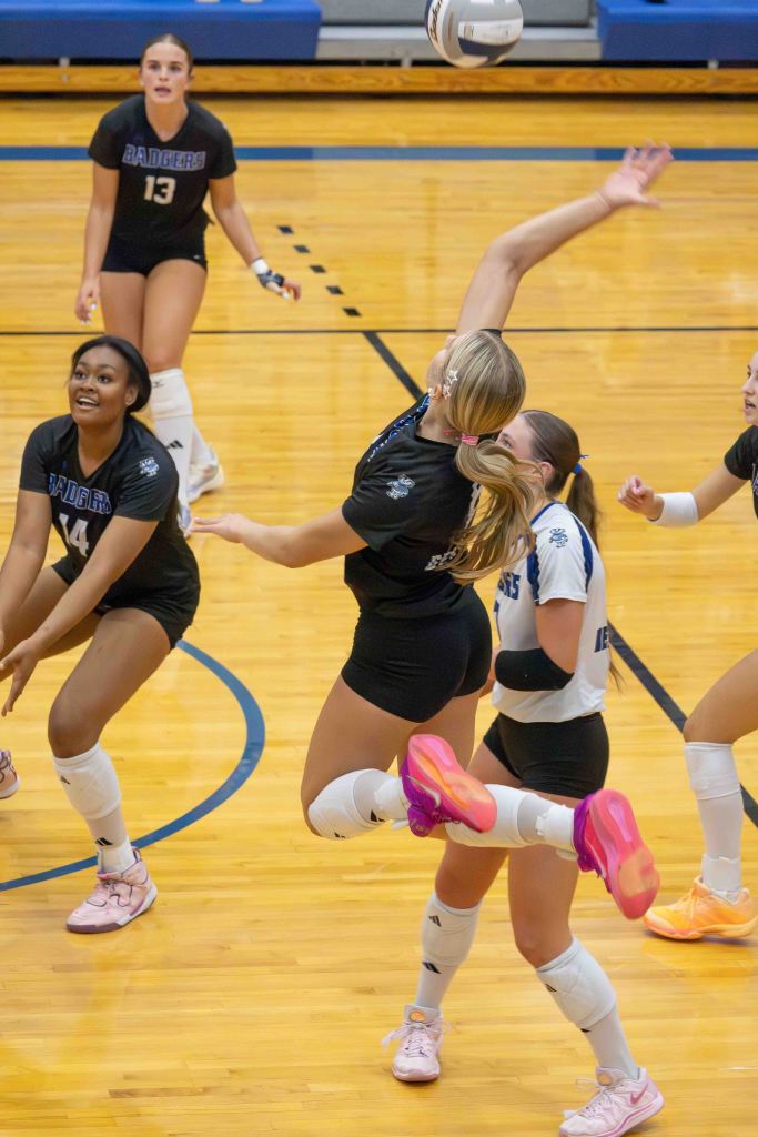 A group of volleyball players in a gymnasium, with one player jumping to strike the ball while others watch and prepare.