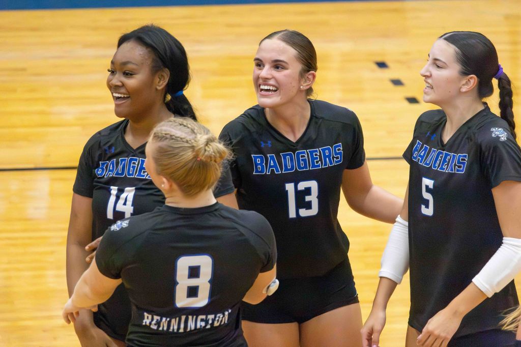 Four female volleyball players celebrating together on the court, wearing matching black jerseys with 'BADGERS' printed on them.