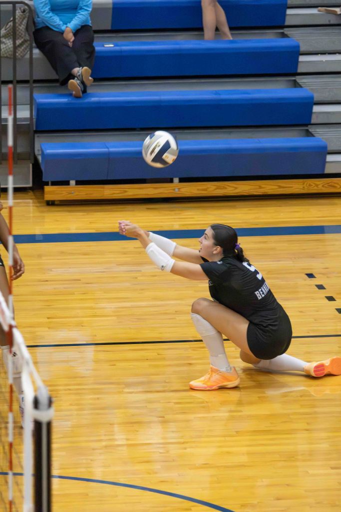 A volleyball player in a black uniform crouches to receive a ball during a game, while spectators sit in the background on blue bleachers.