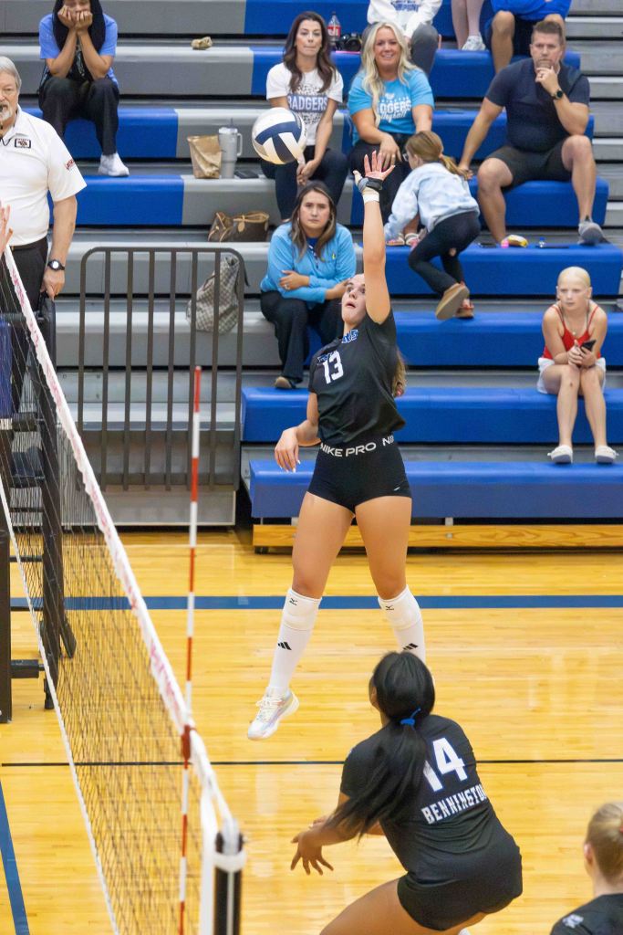 A volleyball player in a black uniform jumps to spike the ball over the net during a game, with spectators in the background.
