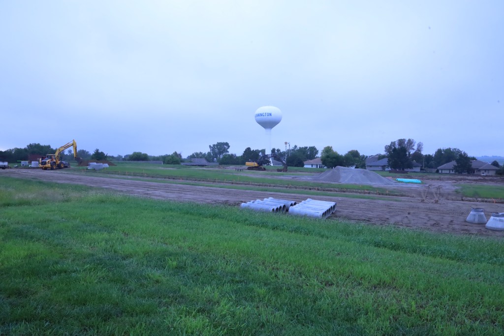 Construction site with heavy machinery, including an excavator and several concrete pipes, set against a background of a water tower labeled 'Simington' and houses, under cloudy skies.