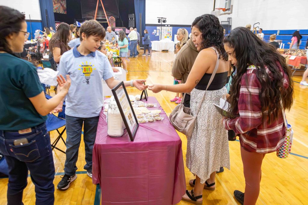 A young boy in a blue t-shirt is presenting a selection of baked goods at a booth to two young women, one wearing a white dress and the other in a plaid shirt. The scene takes place in a gymnasium filled with people and vendor tables.