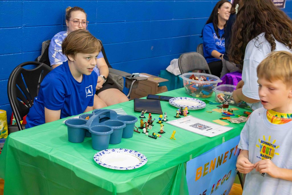 A young boy in a blue shirt sits behind a table covered with a green cloth, displaying various LEGO figures and toys. A child in a white shirt interacts with the display. Several adults are seen in the background, some observing the activity.