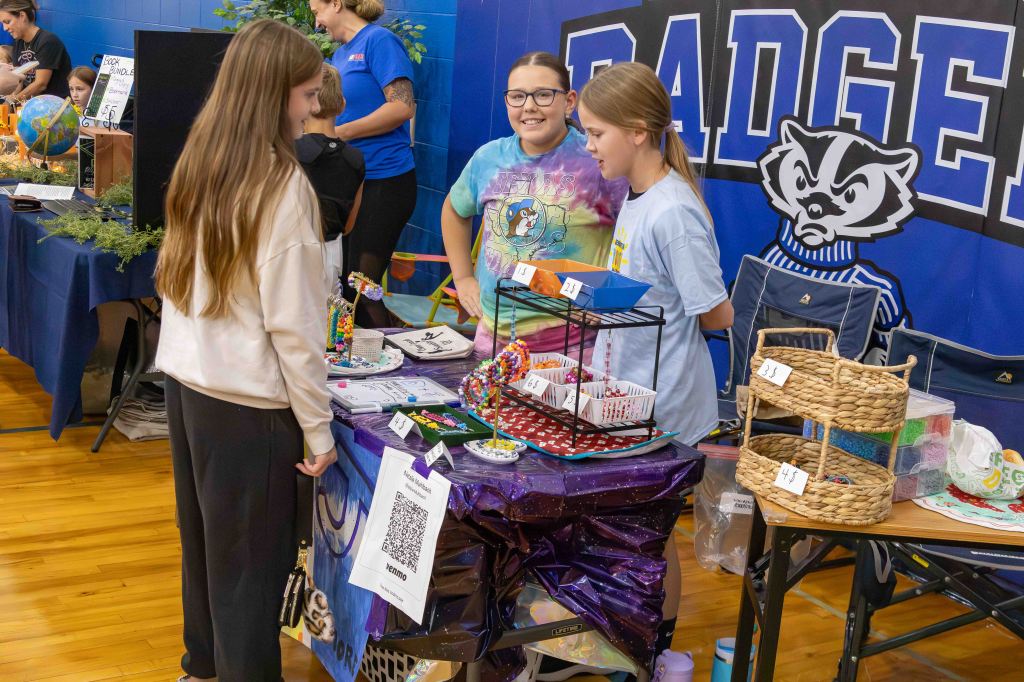 Three girls interacting at a craft vendor table during a school event, showcasing handmade items and colorful crafts.