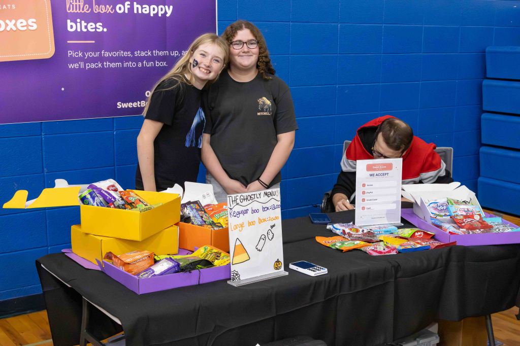 Two smiling girls stand behind a table filled with colorful candy boxes, while a third person is seated, writing something. A sign displays candy options and payment methods accepted.