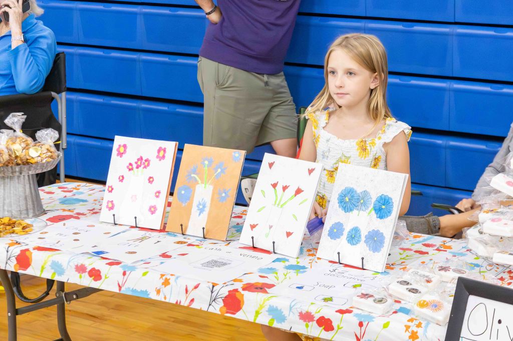 A young girl sitting at a table displaying colorful flower artwork, with baked goods and craft items in a gymnasium setting.