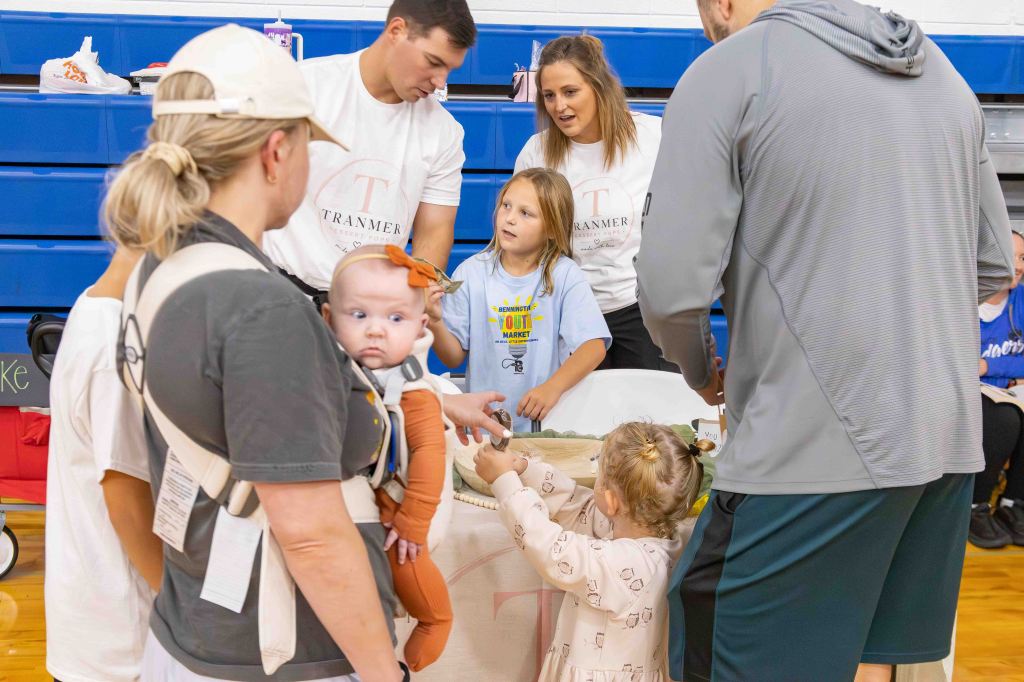 A group of adults and children interacting at an event, featuring a baby being held by a woman, while another child reaches towards a table with various items.