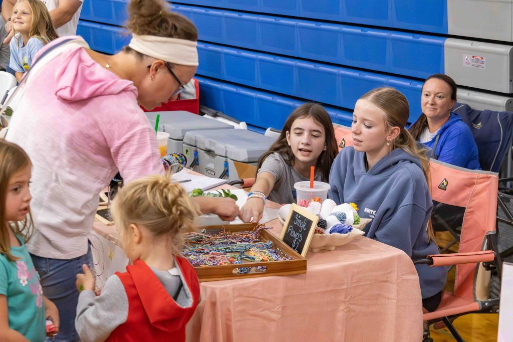 A group of children and adults interact at a craft fair booth, with a table displaying handmade items like bracelets and flowers. One child points to the table while another watches, and an adult stands nearby assisting.