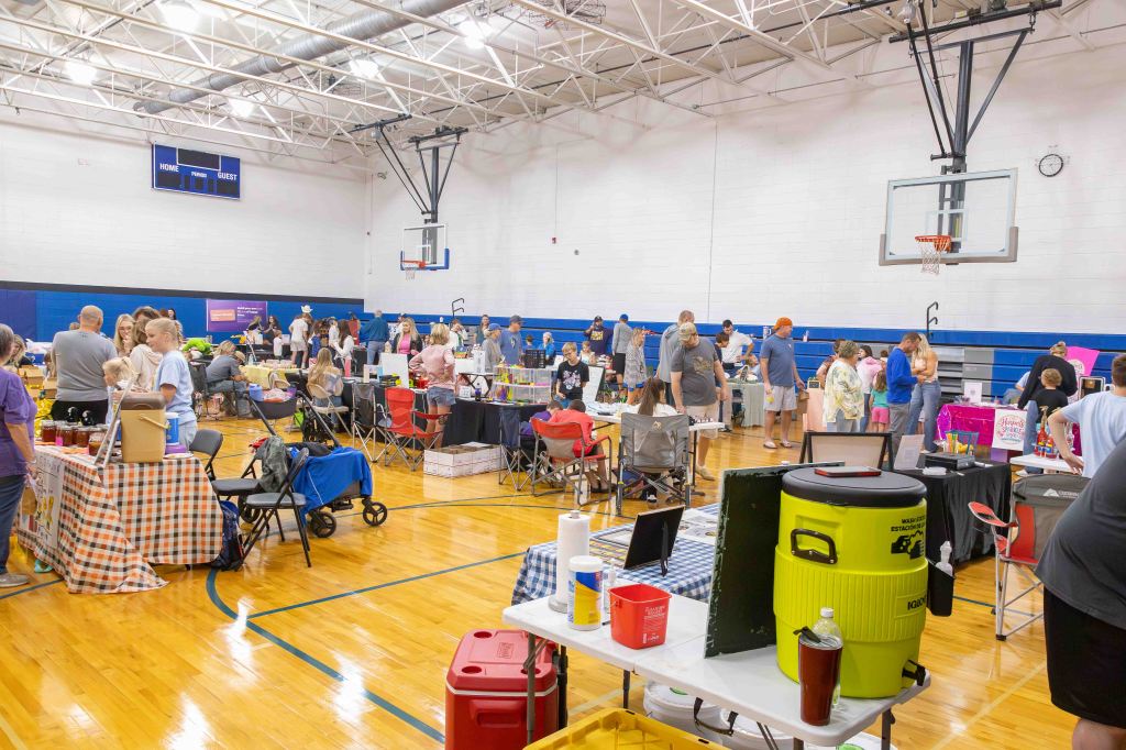 A busy indoor gymnasium filled with people attending a community event, featuring numerous tables displaying various goods and activities. Attendees are mingling and browsing, with colorful signs and products visible.