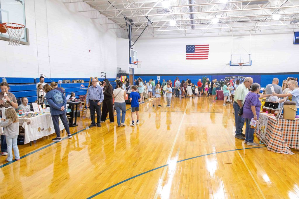 A bustling indoor market scene with people shopping at various vendor booths in a gymnasium. Tables display a variety of products, and attendees engage in conversation while exploring the offerings. An American flag hangs on the wall in the background.