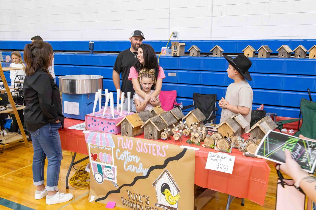 A lively scene at a community fair featuring a booth with cotton candy and handmade crafts. Two sisters pose together, with one girl hugging the other from behind. Various wooden birdhouses and animal figures are displayed on the table, which has a colorful sign reading 'Two Sisters' and a price tag for birdhouses. Attendees engage with the booth and each other in a festive atmosphere.