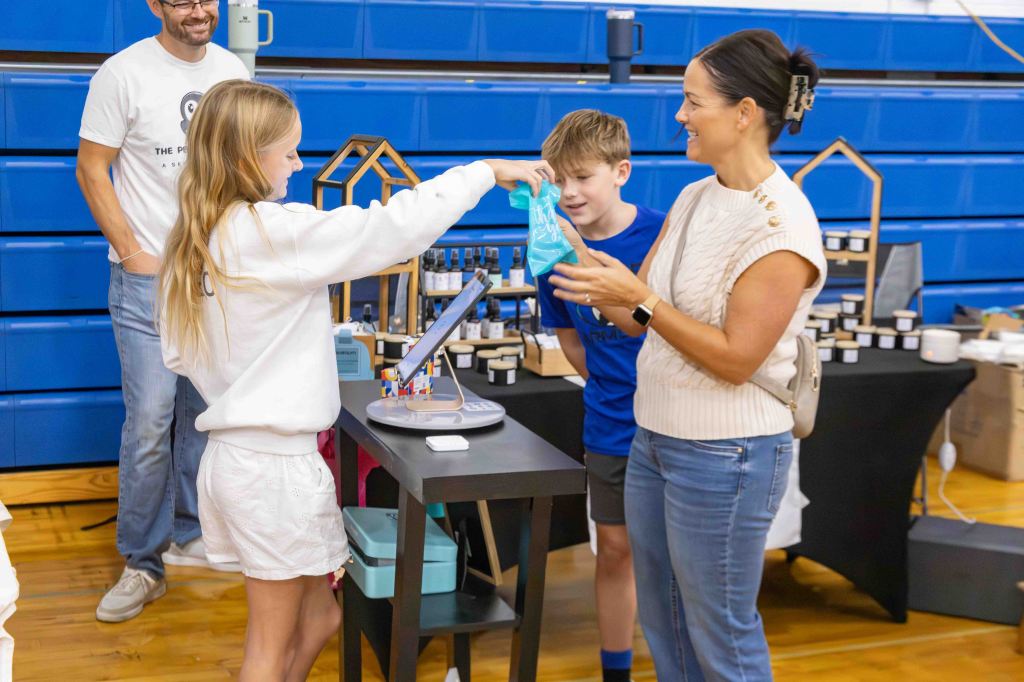 A girl hands a blue bag to a woman at a vendor's table in a gym setting, while a boy watches. A man smiles in the background, and various products are displayed on the table.