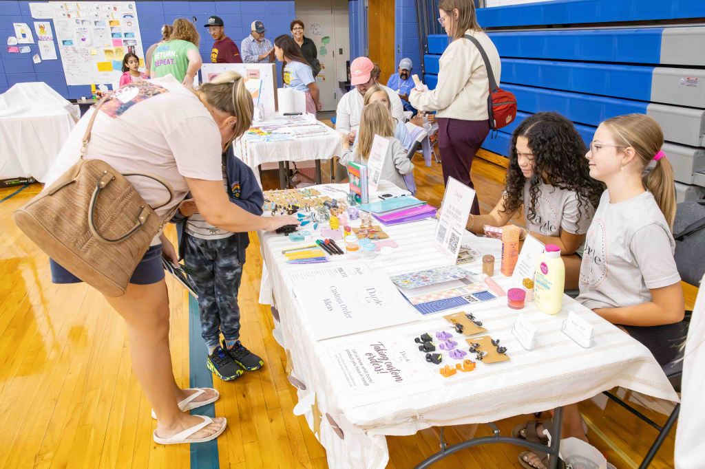 A busy craft fair scene with a table displaying handmade items and several children and adults interacting. One adult is helping a child at the table while others browse in the background.
