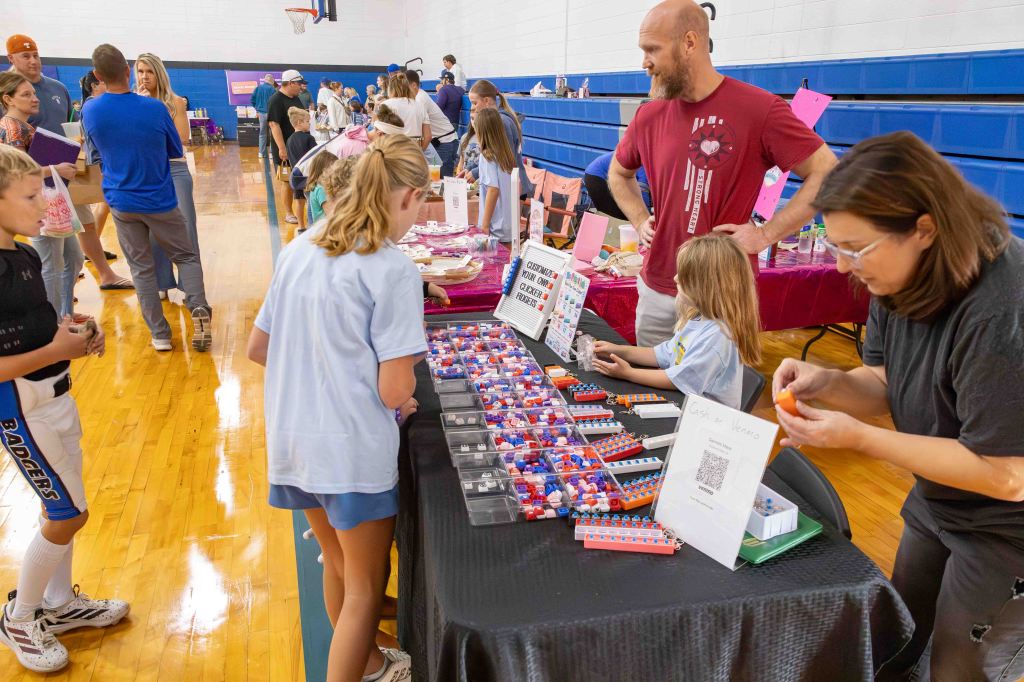 A busy school event featuring children and adults interacting at various vendor tables. Kids are examining colorful items and treats on display, while adults engage in conversation. The scene captures the lively atmosphere of community participation.