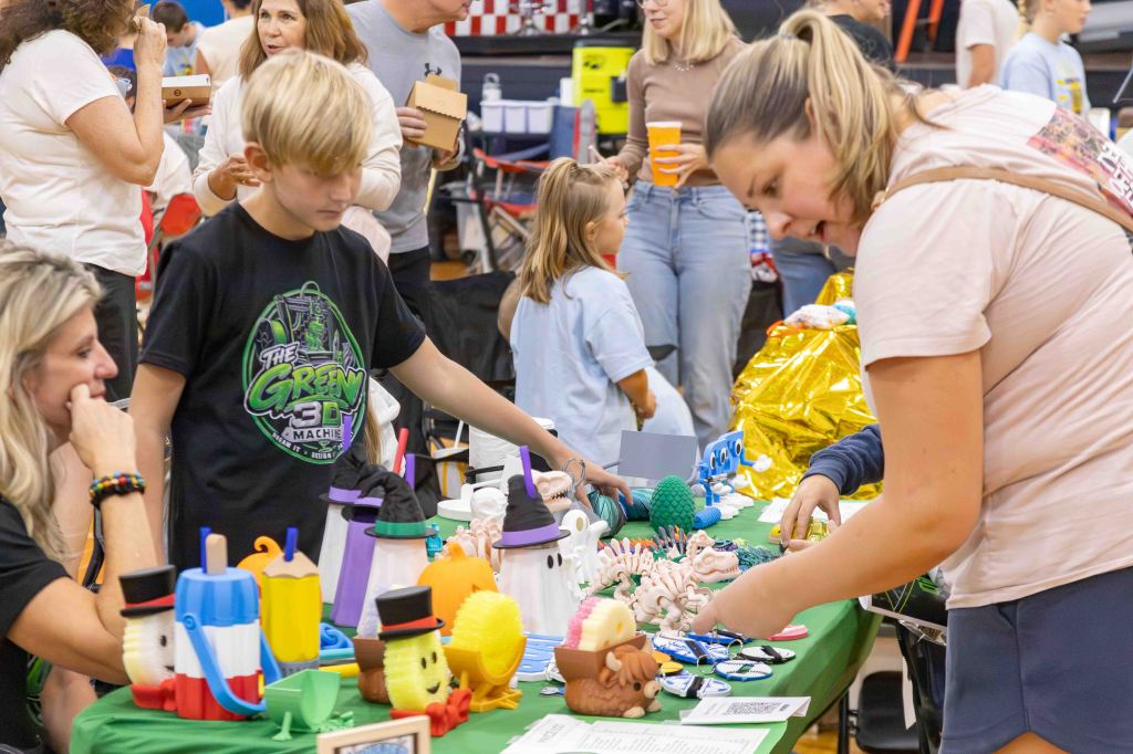 A busy indoor event with people gathered around tables featuring colorful craft items, including decorations and toys. A young boy in a black shirt examines the table, while two women engage in conversation. Children and adults are visible in the background.