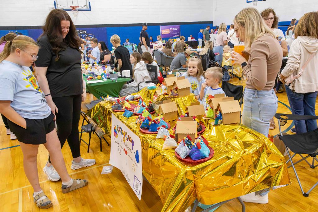 A lively indoor event showcasing children and adults gathered around a colorful craft table with handmade items. People are engaged in conversation and browsing creative displays.