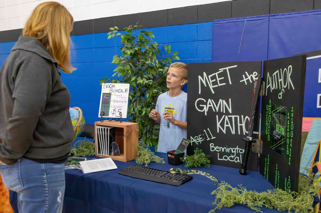 A young boy, Gavin, aged 11, stands at a table promoting his book at an event. He is engaged in conversation with an adult woman who is listening attentively. The table features a 'Book Bundle' sign with items for sale, including a signed copy of his book, bookmark, and stickers. There are decorations, including plants, a globe, and a black display board that highlights Gavin as an author from Bennington Local.