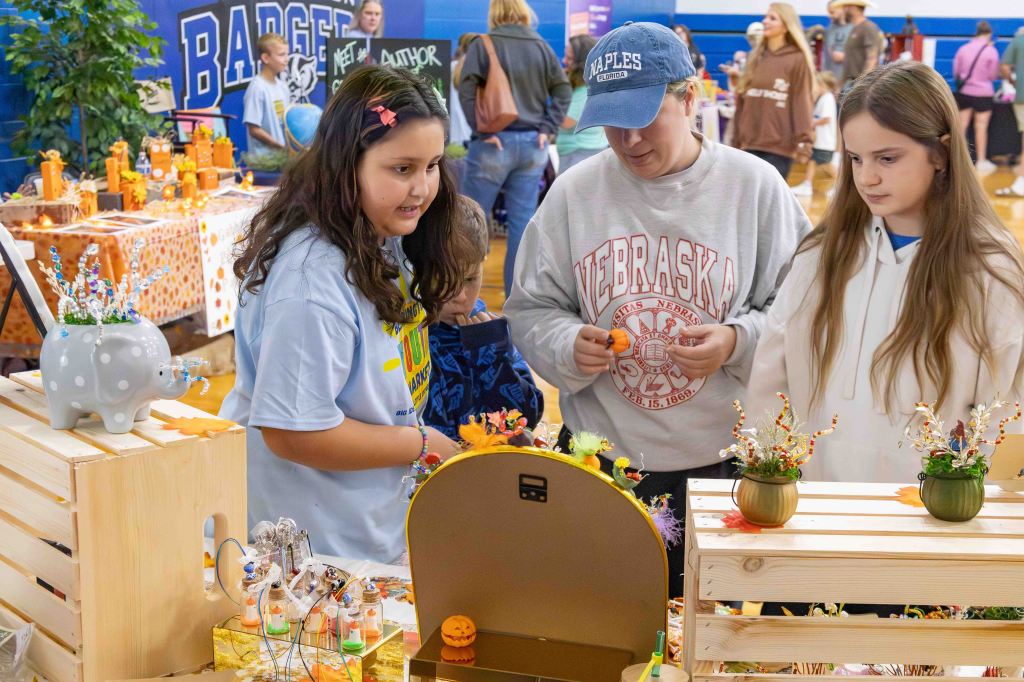 Three children examine crafts at a school fair, with handmade items displayed on tables in the background.