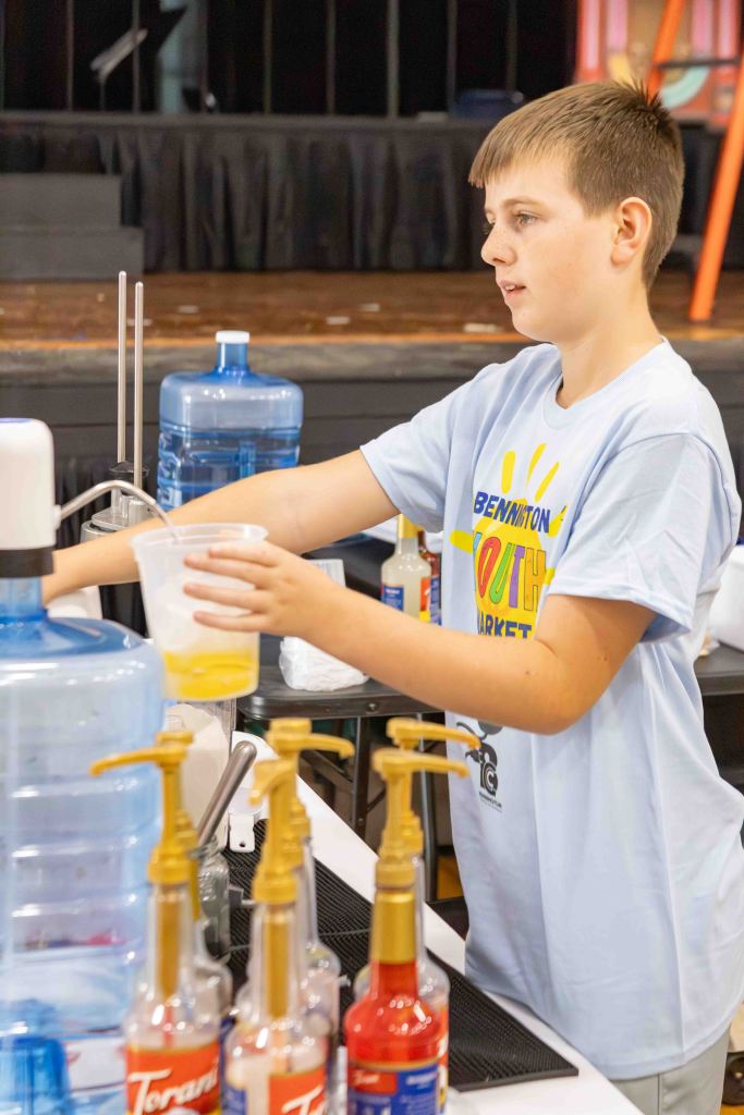 A young boy wearing a light blue t-shirt prepares a drink at a beverage station, handling a cup and pouring from a large water container.