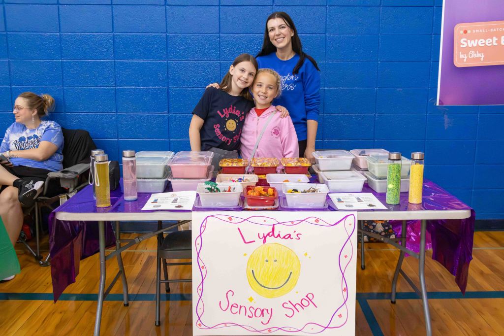 A woman and two girls stand behind a table filled with sensory products, including colorful containers and a sign that reads "Lydia's Sensory Shop." The background features blue walls and a gym setting.