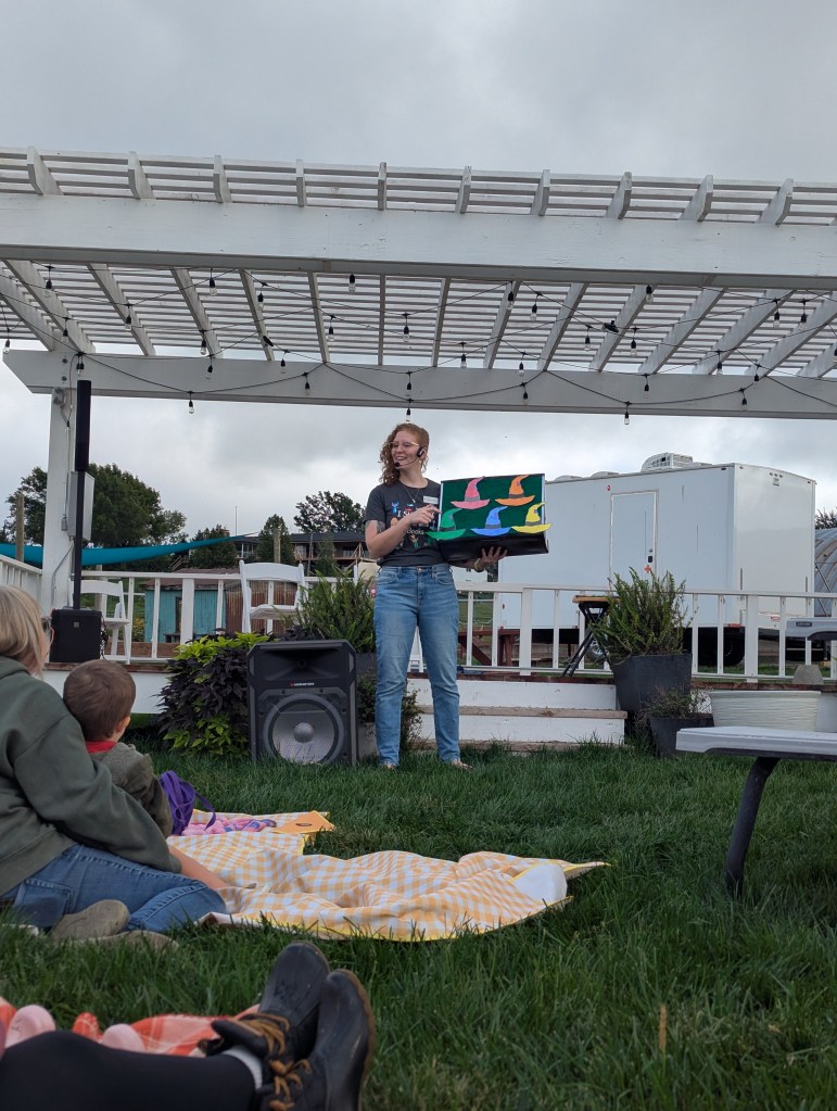 A woman presenting colorful artwork to an audience sitting on the grass under a pergola. The sky is cloudy.