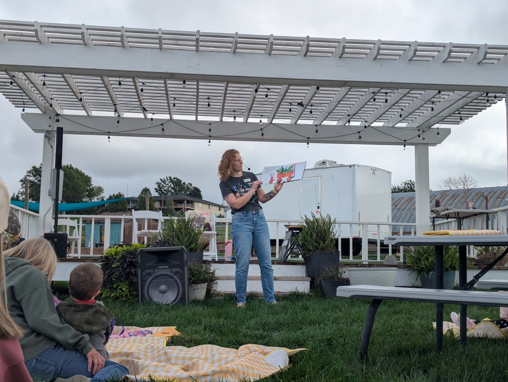 A woman stands on a stage under a pergola, reading from a book to an audience seated on the grass. She is holding a colorful page of the book, with a picnic table and plants in the background.
