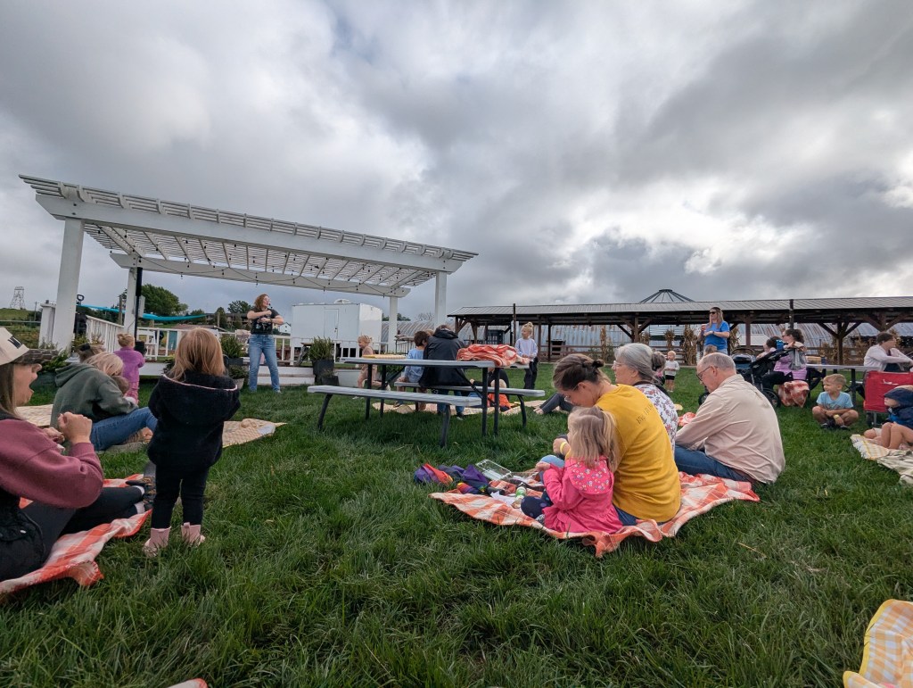A gathering of people sitting on blankets in a grassy area, listening to a speaker at an outdoor event under a pergola, with cloudy skies above.