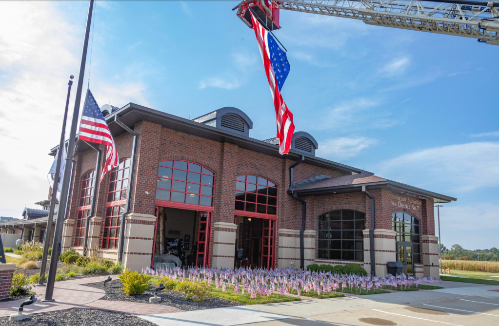 A view of a brick fire station building with large windows, surrounded by small American flags in the foreground and flags on flagpoles, under a clear blue sky.