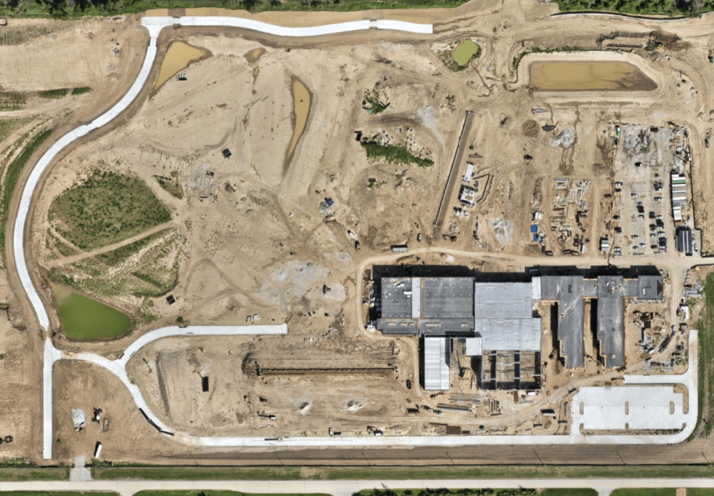 Aerial view of a construction site with a partially built structure, surrounded by dirt and several ponds, with winding concrete paths and construction equipment visible.