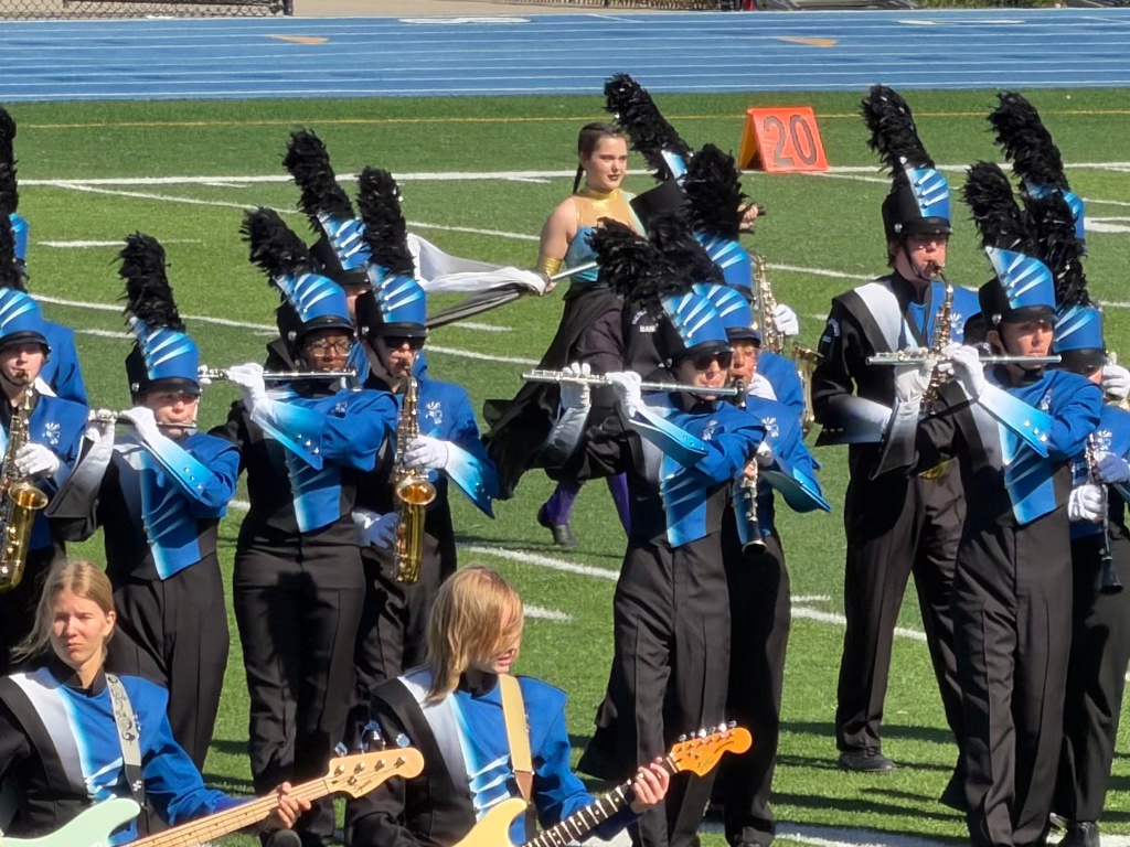 A marching band in blue and black uniforms performs on a sports field, with musicians playing various instruments, including flutes and saxophones. A color guard member in the background waves a flag.