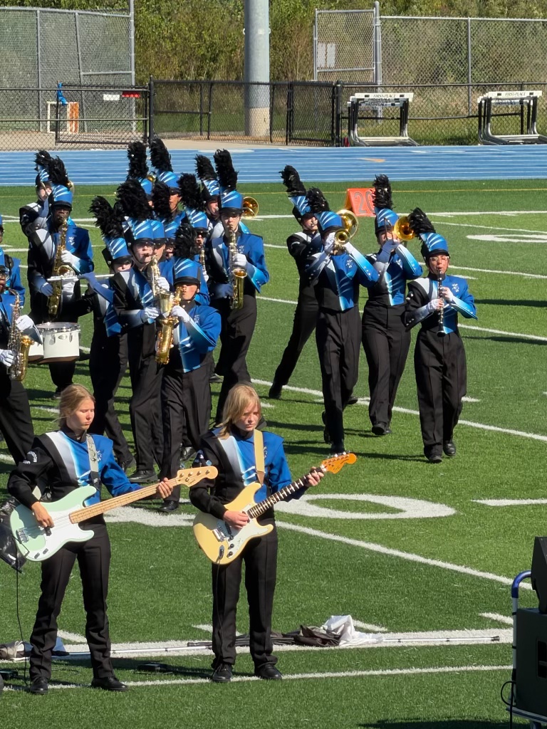A marching band performing on a sports field, featuring musicians in blue and black uniforms. Two guitarists in the foreground play electric guitars while the rest of the band is positioned behind them, playing brass instruments.