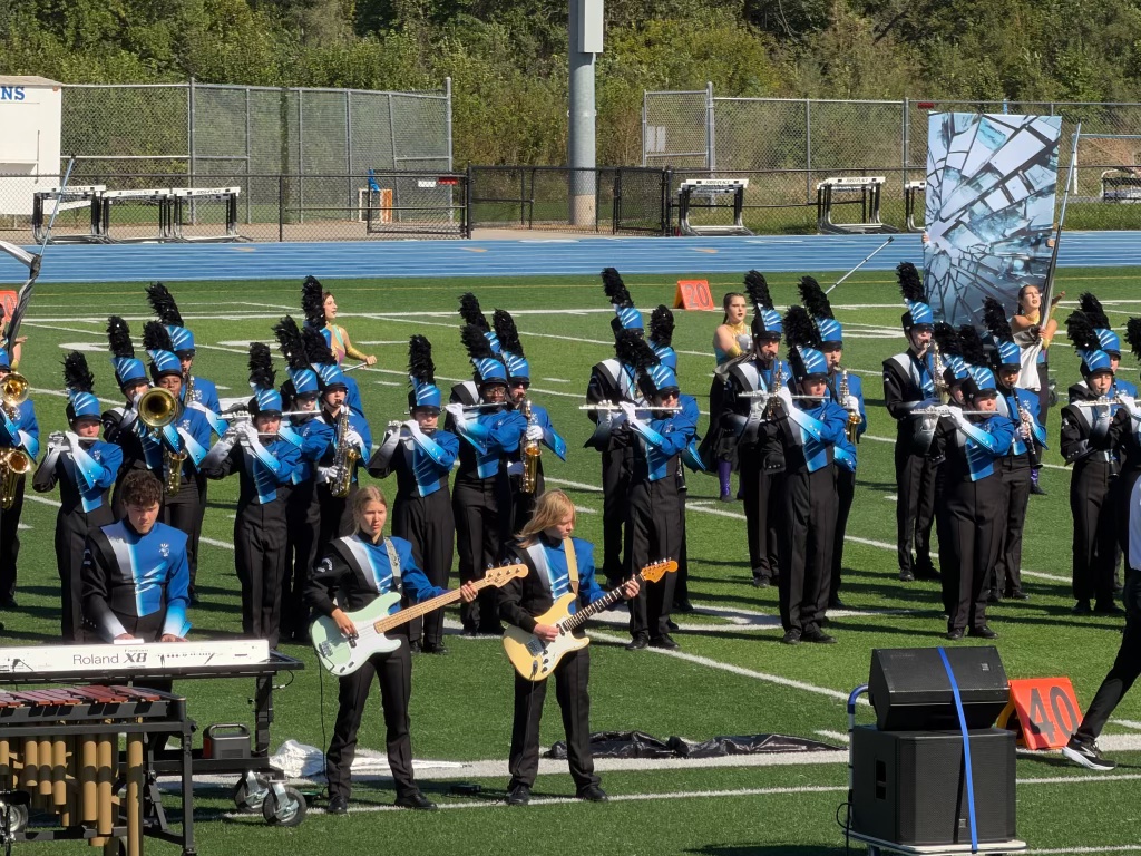 A marching band performing on a green football field, featuring musicians in black and blue uniforms, with brass instruments and percussion equipment. Some members are playing electric guitars and keyboards, while colorful flags are being displayed.