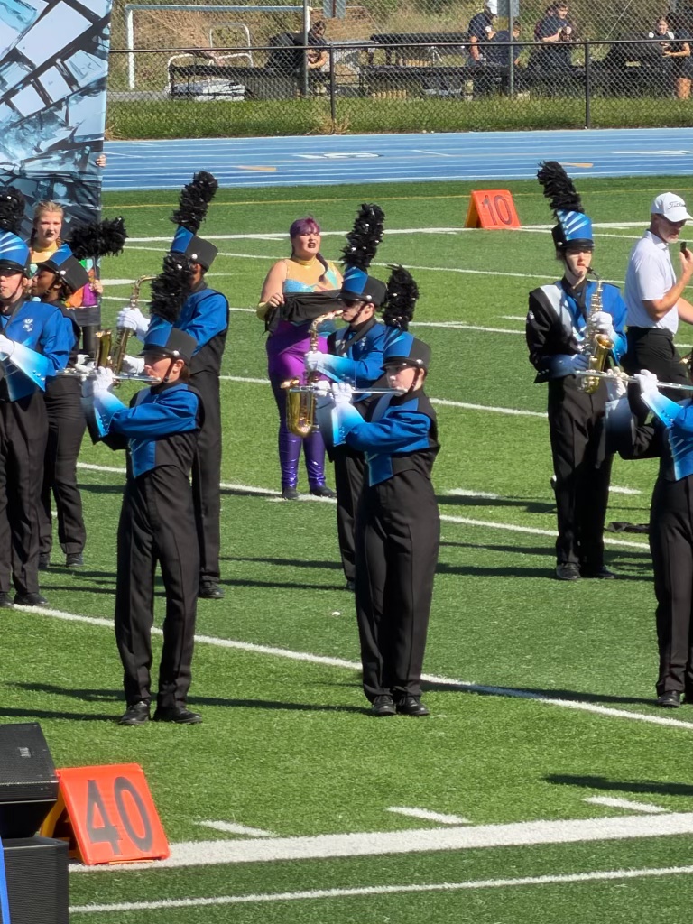Marching band performing on a football field, with members in blue and black uniforms, holding instruments, and a banner in the background. A singer in colorful attire is visible behind the band.